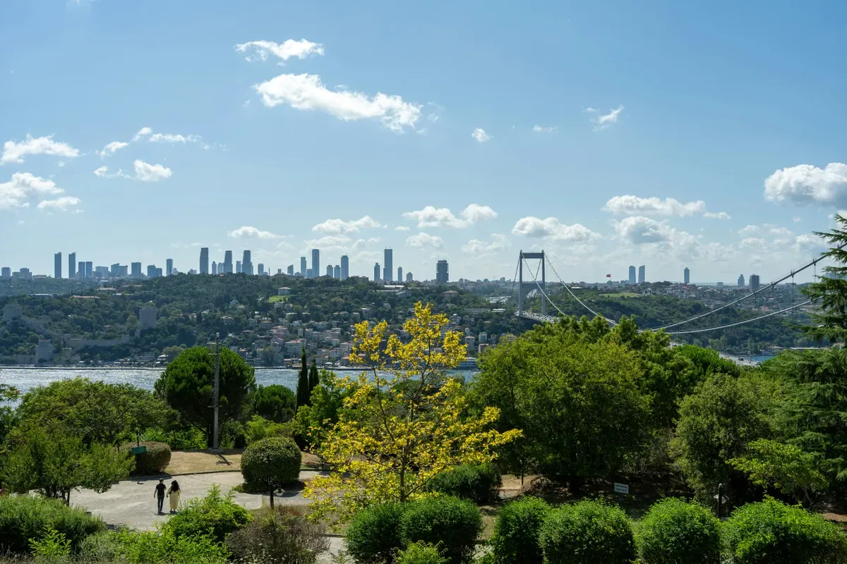 Un parco di Istanbul con alberi e aiuole, con lo skyline e il ponte sul Bosforo sullo sfondo