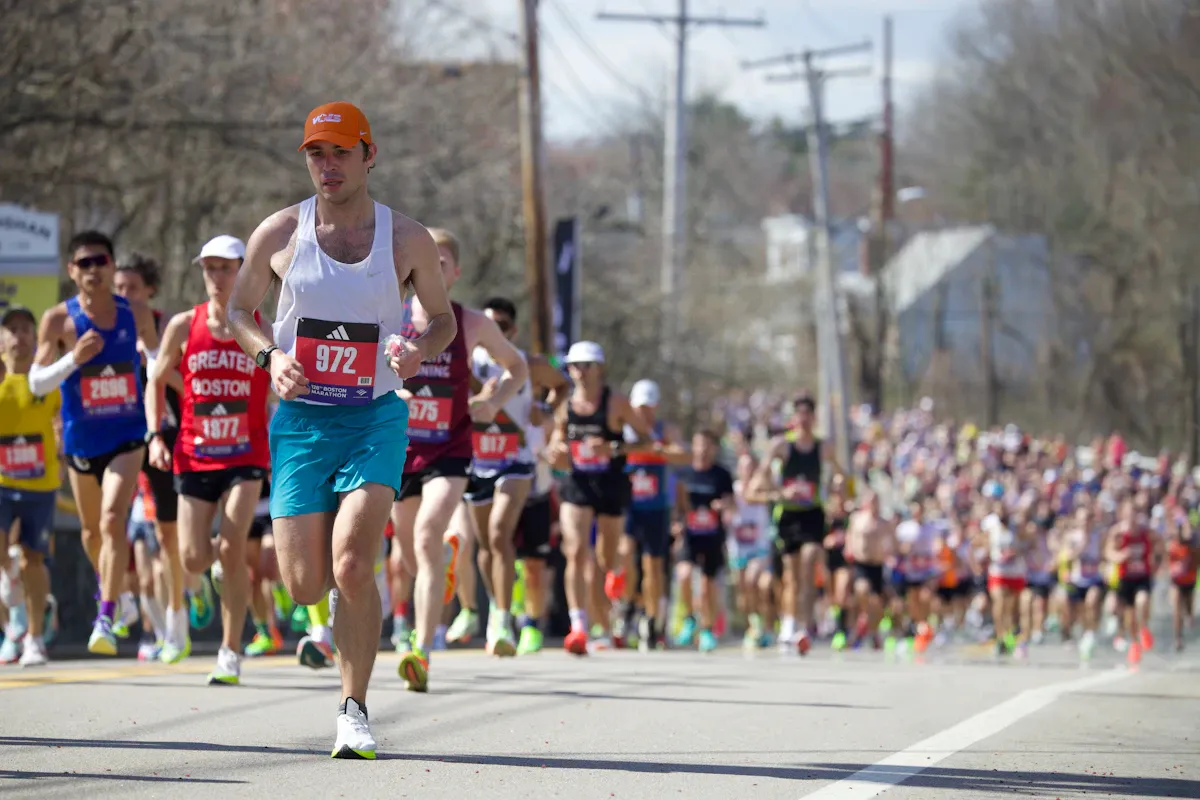 Un runner con il cappellino arancione in testa a un gruppo lungo una strada alberata della maratona di Boston