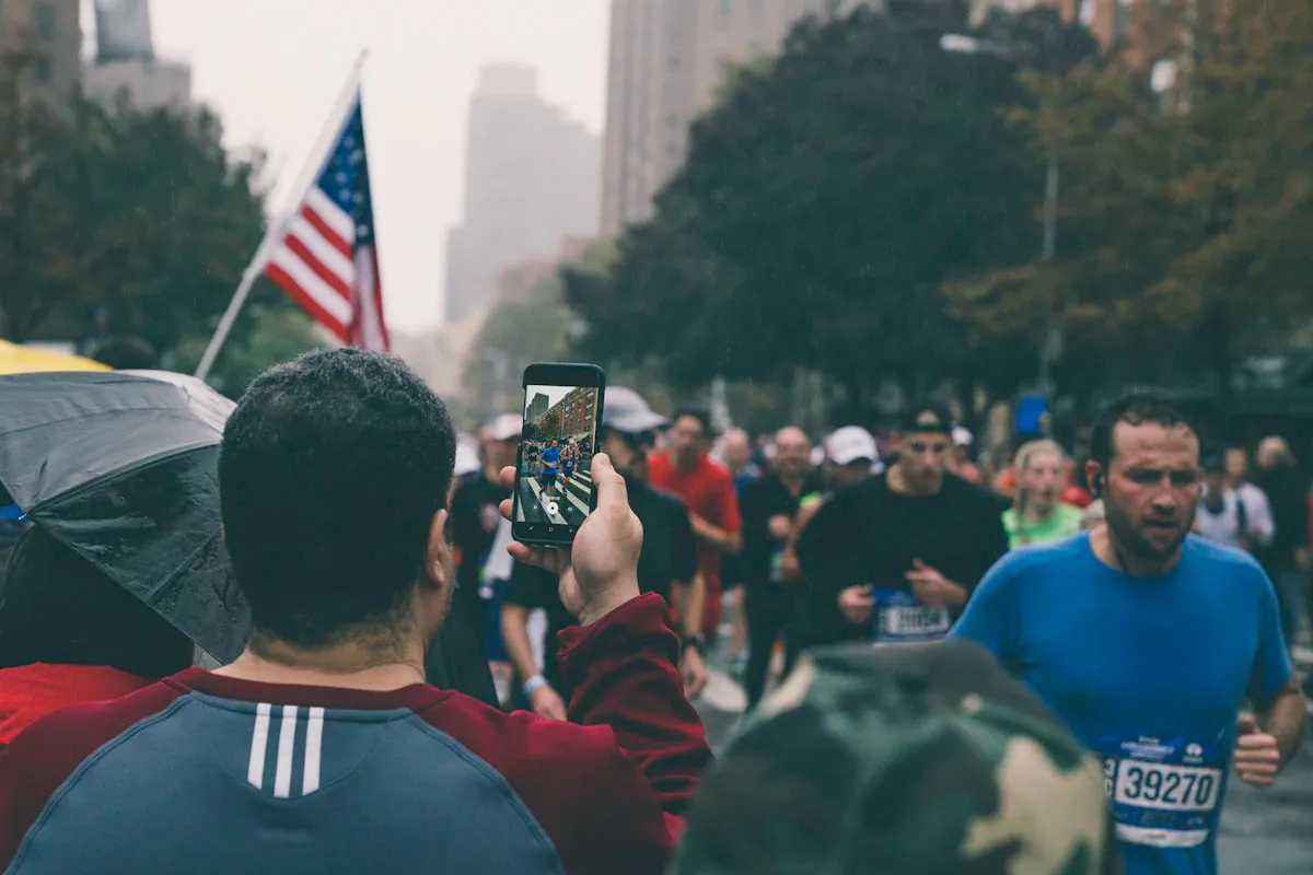 Uno spettatore riprende con il telefono i runner della maratona di New York, con la bandiera americana sullo sfondo